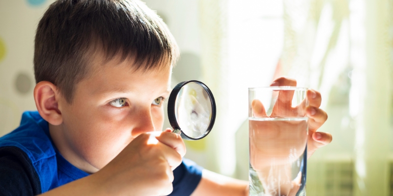 Young boy in a blue shirt uses a magnifying glass to examine a glass of water.